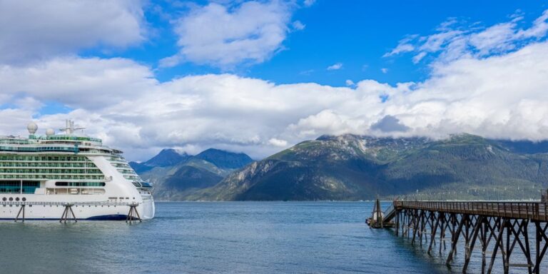 A white cruise ship docked near a wooden pier with a backdrop of towering mountains under a partly cloudy sky. The serene waters reflect the grandeur of the Alaskan landscape, emphasizing the calmness of the cruise experience.