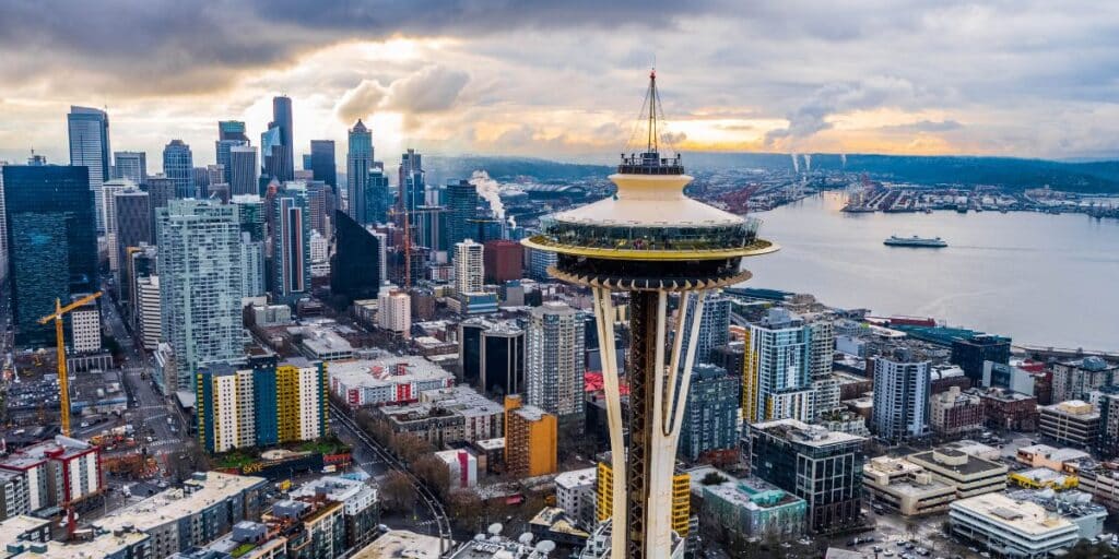 Aerial view of Seattle skyline featuring the iconic Space Needle with a cloudy sky backdrop, foreground showing dense urban architecture with various construction cranes, and Elliott Bay in the distance.