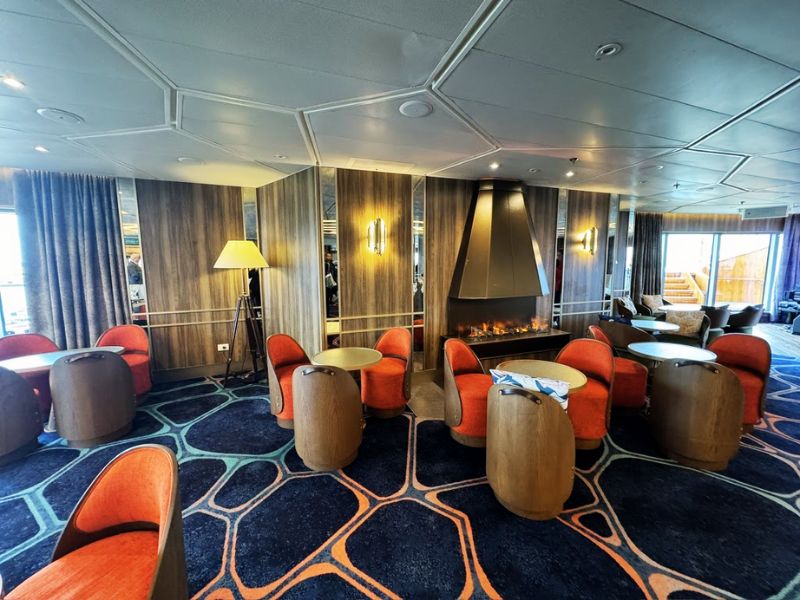 The Club on the Seabourn Venture, which is styled as a chic and cozy lounge area perfect for relaxation and social gatherings. The space features modern, comfortable seating in vibrant orange and neutral tones, complemented by stylish, barrel-shaped wooden tables. A prominent feature is the elegant, central fireplace, adding a warm and inviting ambiance. The design is completed with a dynamic, patterned carpet and wood paneling, creating a sophisticated yet welcoming environment for guests to enjoy leisure time or evening cocktails.