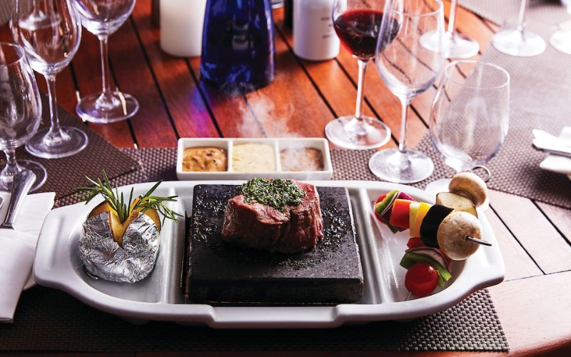 A gourmet meal at The Grill on a Silversea cruise ship, showcasing a succulent steak topped with herbs on a hot stone, accompanied by a skewer of grilled vegetables and a selection of sauces, with elegant glassware and wine bottles in the background, inviting an exquisite dining experience.