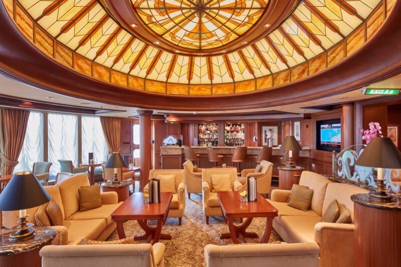 Inside the Grills Lounge on Cunard's Queen Elizabeth, featuring a warm and inviting atmosphere with a stunning stained-glass ceiling dome. Plush beige armchairs and sofas are neatly arranged around wooden coffee tables, with elegant lamps providing soft lighting. In the background, a well-stocked bar awaits, enhancing the cozy yet refined ambiance of this exclusive lounge area.