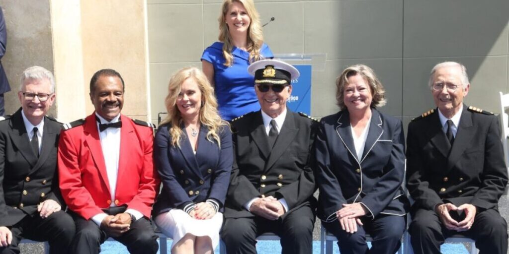 The Love Boat Cast posing together at a Princess Cruises event. The men are dressed in naval-style uniforms and the women in business attire, with one in a casual blue top. They are smiling and seated in front of a podium with a clear sky in the background.