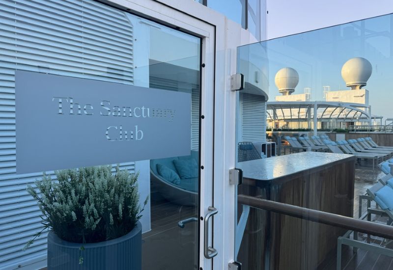 The image shows the entrance to The Sanctuary Club on Princess Cruises' Sun Princess. The entrance features a glass door with "The Sanctuary Club" elegantly etched into the glass. Inside, you can see part of a luxurious outdoor lounge area with comfortable seating and loungers, designed for relaxation in a serene and exclusive environment. The area is well-appointed with stylish furniture and is partially sheltered, with additional seating and amenities visible beyond the door. The backdrop includes the ship's upper deck with radar domes, further emphasizing the premium and secluded nature of this space.