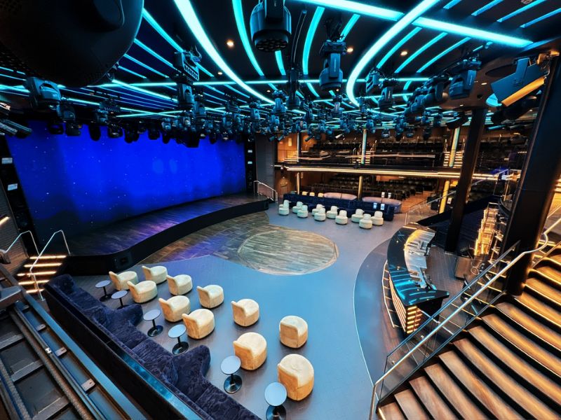 The modern and stylish theatre on Costa Smeralda, featuring a spacious stage with dynamic blue lighting, comfortable seating areas with plush sofas and cube stools, and state-of-the-art audio-visual equipment ready for a performance.