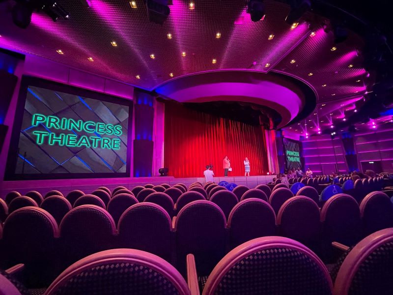 Interior view of the Princess Theatre on a Princess cruise ship, showcasing a sea of purple seats facing a large stage with a red curtain and a screen displaying 'Princess Theatre'. The ceiling is dotted with lights, enhancing the venue's vibrant atmosphere.