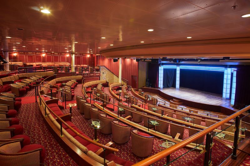 The grand theater on a Silversea cruise ship, featuring rows of plush red seats, elegant wooden balustrades, and a large stage set for performance, all under warm ambient lighting that creates an inviting space for entertainment at sea.