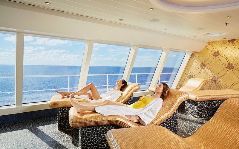 Two guests relax in the serene Thermal Suite on a Carnival cruise ship, reclining on contoured loungers that offer ocean views through panoramic windows. The space is designed for tranquility, with a mosaic-tiled floor and golden accents creating a luxurious spa experience at sea.