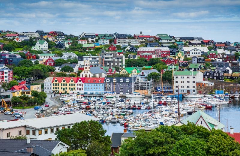 This image beautifully captures the vibrant and colorful town of Torshavn, the capital of the Faroe Islands. The town is densely populated with picturesque houses painted in various bright colors, and their roofs are distinctly red, green, and blue. The bustling harbor is filled with boats, suggesting a strong local maritime culture. Lush greenery surrounds the area, and the town's architecture, combined with its natural setting, presents a charming and inviting atmosphere characteristic of this North Atlantic island community.