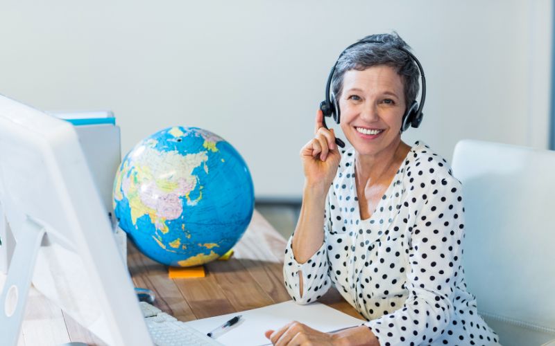 A smiling travel agent wearing a headset sits at her desk with a large colorful globe beside her, signaling readiness to assist with travel plans, depicted in a bright, modern office setting.