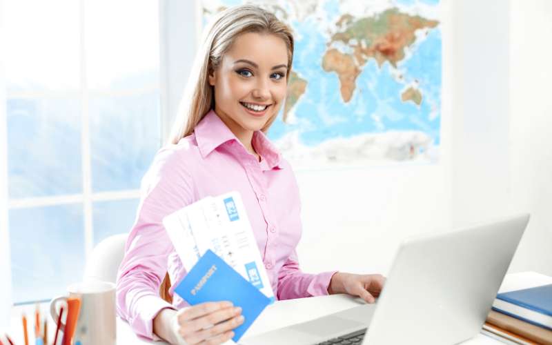 A lady wearing a pink blouse, sat at her laptop passing over a passport and tickets for travelling.