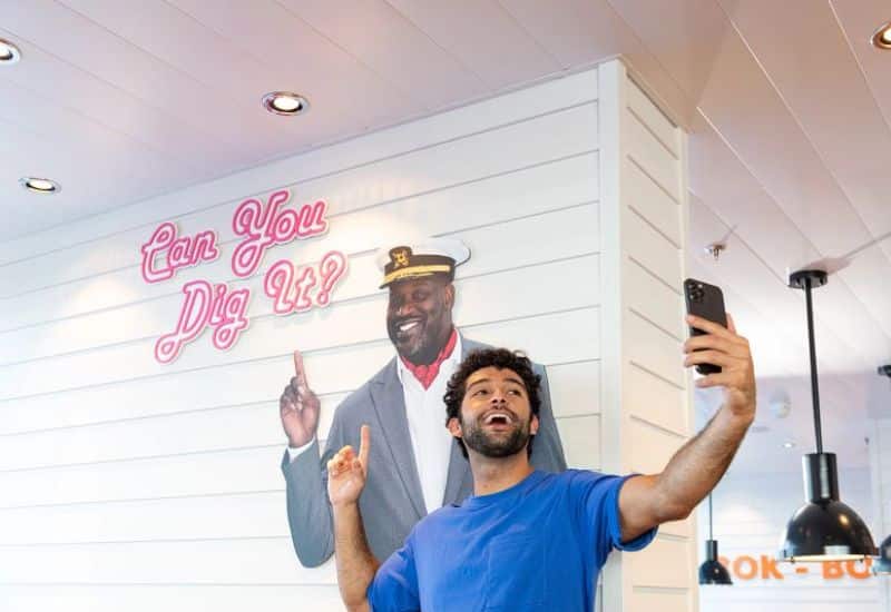 A man in a blue shirt taking a selfie with a playful neon sign that reads "Can You Dig It?" and a mural of a smiling man in a captain's hat pointing upwards.
