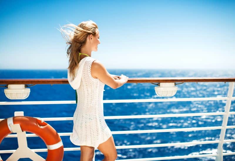 A woman in a white summer dress leaning on the railing of a cruise ship, enjoying the ocean view with her hair blowing in the sea breeze.