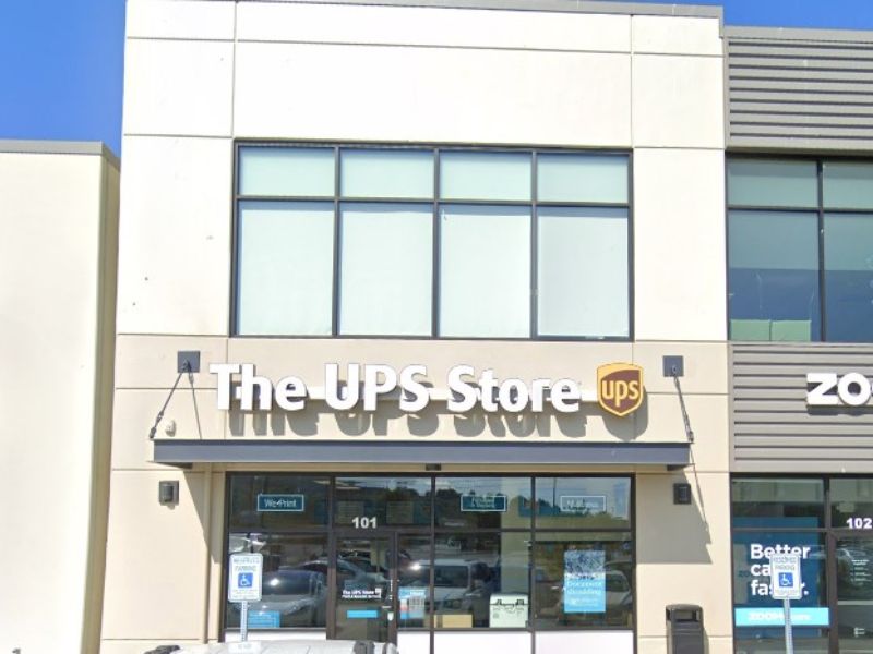 The exterior of The UPS Store with large storefront windows and the distinctive UPS logo, situated in a modern strip mall on a sunny day, showcasing its location for shipping, printing, and business services.