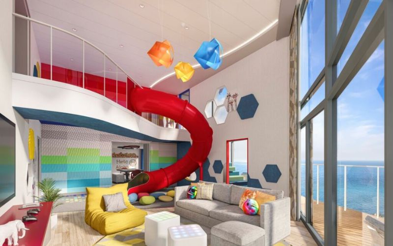 Playful and colorful interior of a family suite on a cruise ship, featuring a bright red slide connecting two floors. The room is bathed in natural light from floor-to-ceiling windows that offer a view of the ocean, with fun decor including oversized candy-like wall ornaments and cozy, contemporary furnishings.