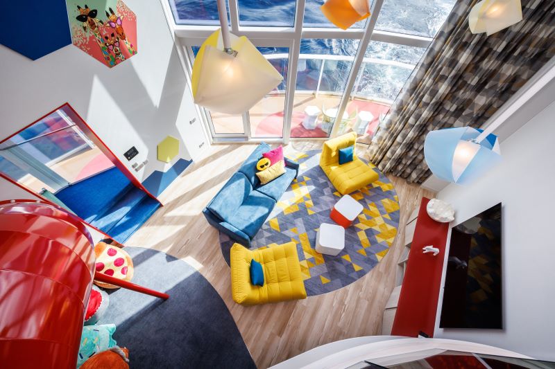 Overhead view of Royal Caribbean's Ultimate Family Townhouse play area, vibrant with colorful decor. The room features a whimsical design with bright yellow and blue furniture, a red slide, funky light fixtures, and a floor-to-ceiling window overlooking the sea. It's a playful and inviting space for children, with various toys and games, fostering a fun and imaginative onboard experience.