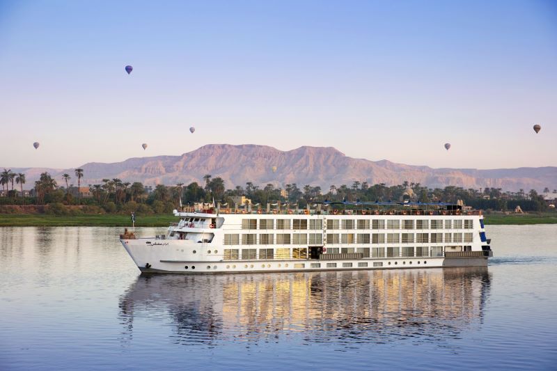 A Uniworld Boutique River Cruise ship serenely sails on the Nile at dawn, with the soft light of sunrise illuminating the desert mountains in the background. Hot air balloons float gracefully in the sky, adding a touch of adventure to the peaceful scene. The ship’s decks are lined with passengers likely taking in the majestic views, mirroring the tranquil and exploratory spirit of a river cruise in Egypt.