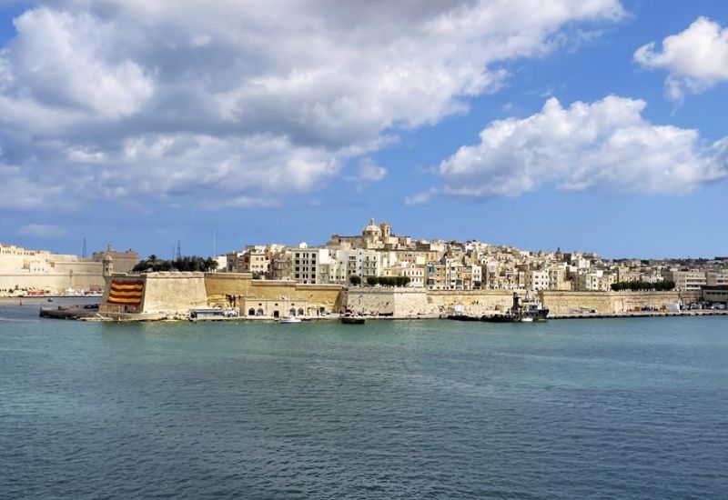 The scenic view of Valletta, the capital city of Malta, as seen from the sea. The city's skyline is characterized by timeless architecture, including sandstone buildings and historical landmarks that rise steeply from the waterfront. Prominent fortifications and walls line the harbor, reflecting Malta's rich history in maritime defense. The serene Mediterranean Sea complements the bustling cityscape under a partly cloudy sky, enhancing the picturesque and inviting atmosphere of Valletta.