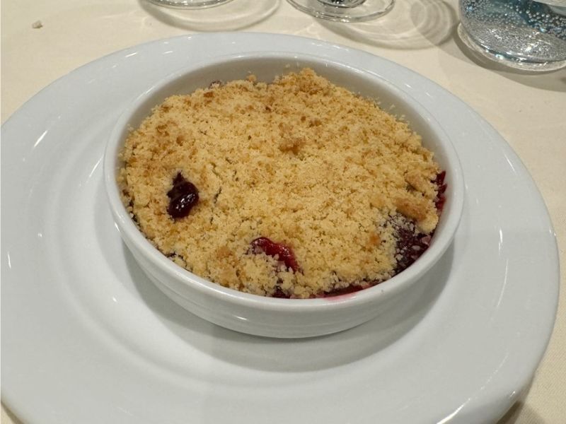 A warm vegan berry crumble dessert in a white ramekin on a matching plate, with a golden brown crumb topping hinting at the juicy berries beneath.