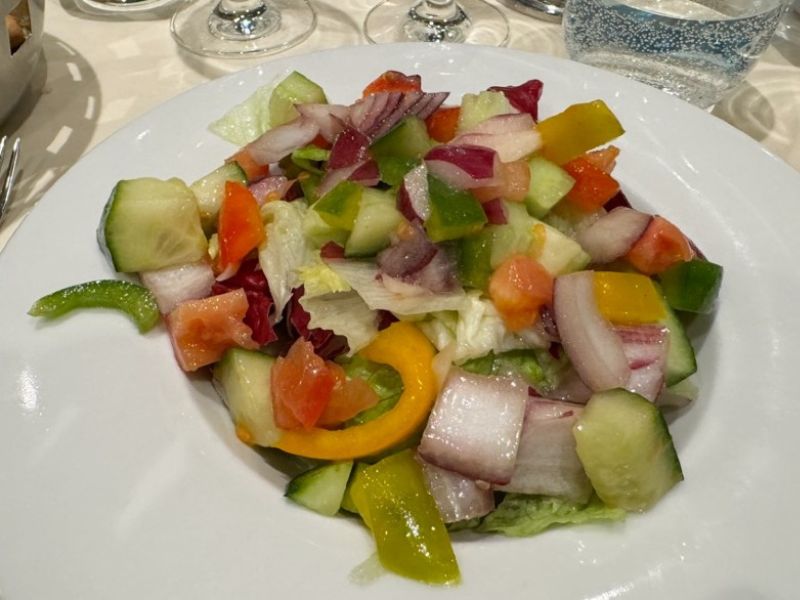 A colorful vegan salad on a white plate, featuring crisp cucumbers, ripe tomatoes, bell peppers, and red onions, served as a fresh starter on MSC Cruises. The salad offers a healthful dining option against the elegant backdrop of a set dining table.