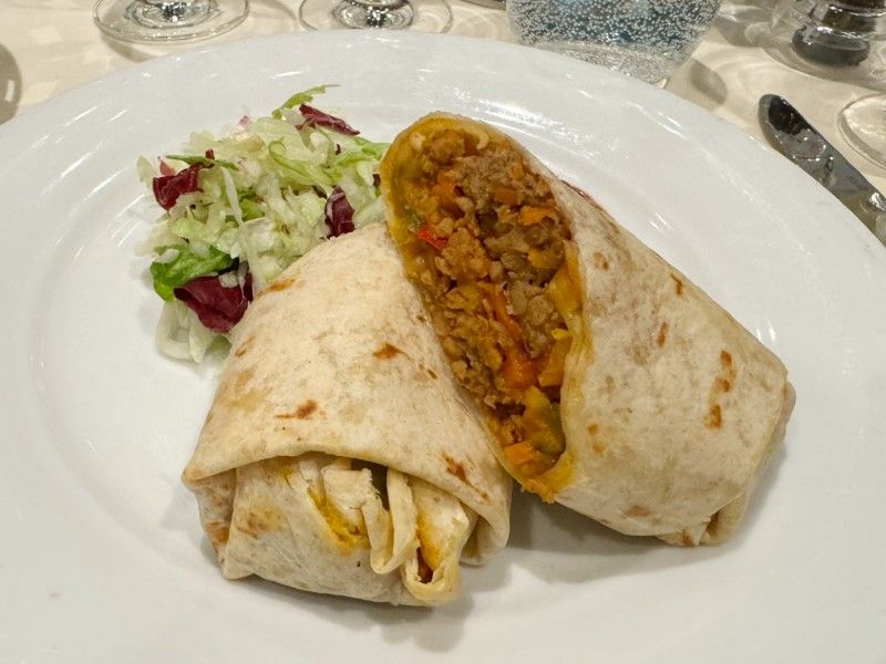 A vegan tortilla wrap filled with a colorful mixture of vegetables and grains, cut in half to showcase the filling, accompanied by a side of crisp mixed salad, presented on a white plate.