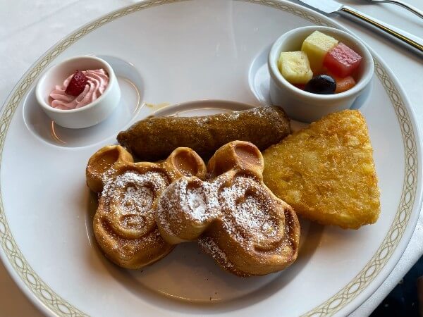 A photo of Mickey Mouse waffles on a plate with fruit and a hash brown.