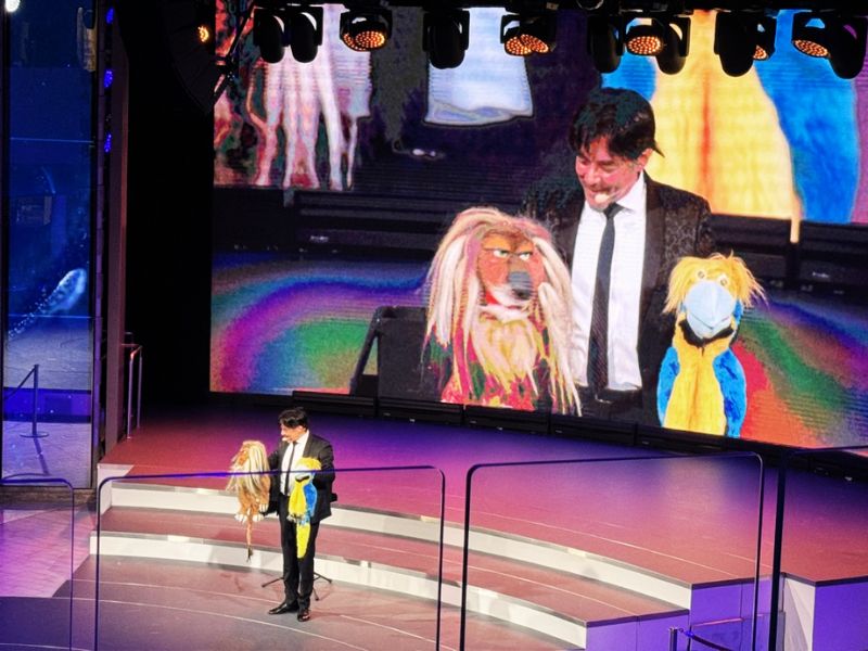 A ventriloquist performing on stage with colorful puppets on Costa Smeralda, engaging the audience with his act, as highlighted by the large screen in the background displaying the show for all to see.