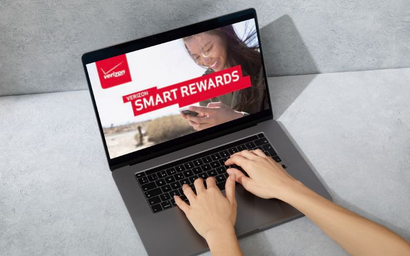 A person is using a laptop displaying the 'Verizon Smart Rewards' program on the screen, featuring a happy customer using a smartphone. The setting suggests a casual environment, perhaps a home or café, with a focus on the benefits of the rewards program.