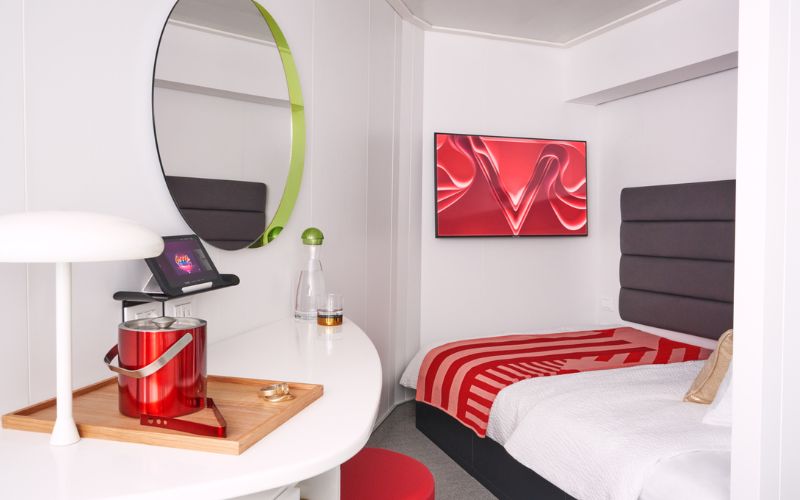 A stylish Solo Sea View cabin on a Virgin Voyages cruise ship, featuring a cozy bed with red and white bedding, and a padded headboard. The room includes a sleek desk area with a modern lamp, a round mirror, and a mounted flat-screen TV displaying vibrant artwork. The contemporary decor is accented with pops of color, creating a chic and inviting space.