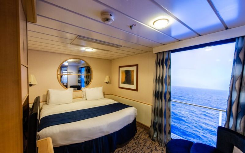 Interior of a cruise ship stateroom with a large bed centered between nautical-themed curtains and a round porthole mirror. The room has a cozy ambiance with a view of the open sea through the virtual balcony