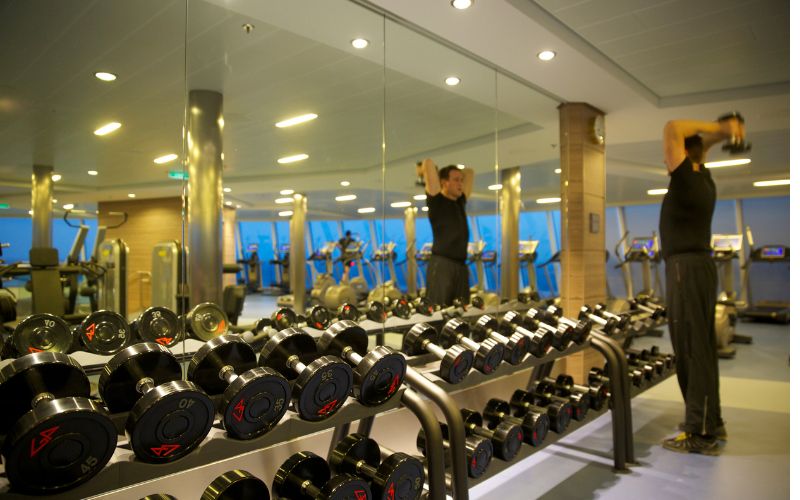 A person lifts weights in a well-equipped gym with rows of dumbbells in the foreground and reflections of exercise machines in the mirrored wall, highlighting an active fitness environment.