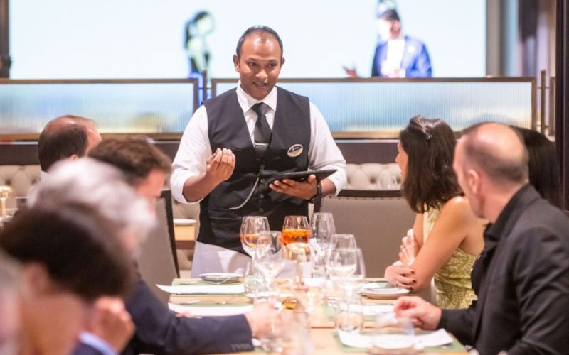 A smiling MSC waiter in a vest and tie taking an order from guests at a well-appointed dining table, with a focus on customer interaction in the main dining room of an MSC Cruises ship.