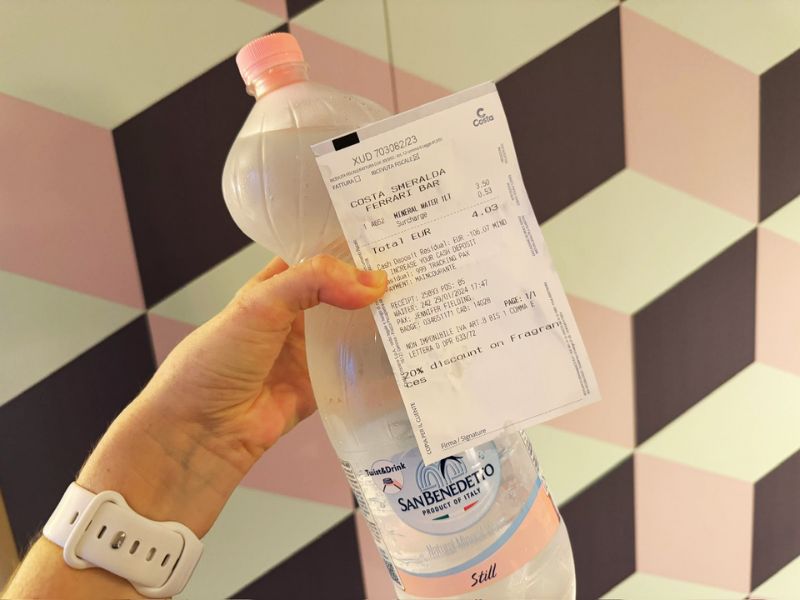 Cruise Mummy holding a transparent bottle of San Benedetto still water with a receipt attached, indicating a purchase onboard the Costa Smeralda, against a geometric-patterned backdrop.