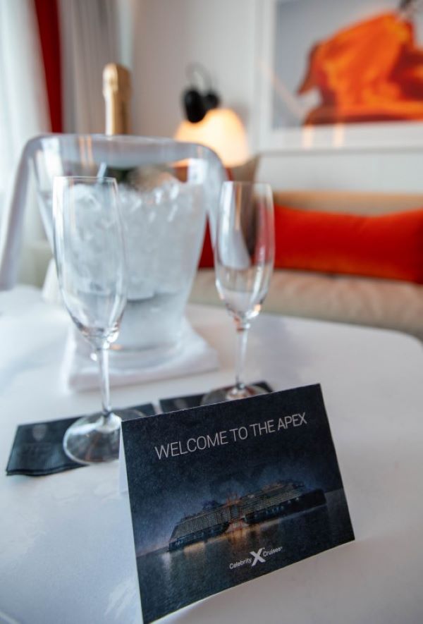 A welcoming setting on Celebrity Apex featuring a chilled bottle of champagne in an ice bucket, two empty glasses ready for a toast, and a 'Welcome to the Apex' card on a table with a vibrant orange sofa and warm lighting in the background.