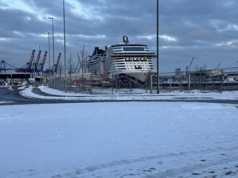 MSC Euribia cruise ship docked at Hamburg cruise port during winter, with a snow-covered ground in the foreground and industrial cranes in the background under a cloudy sky. The image conveys a cold, off-season atmosphere possibly contributing to lower cruise prices.