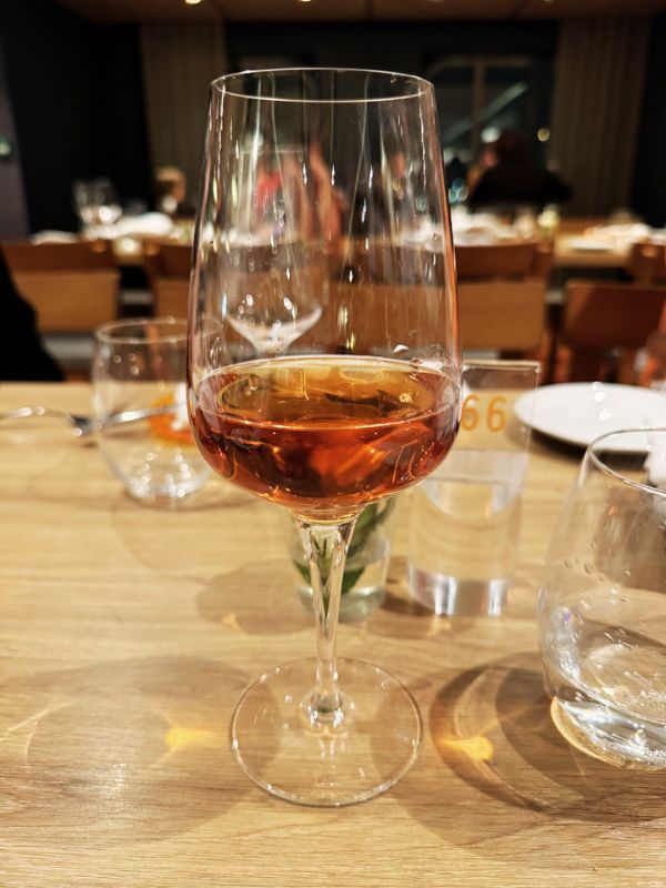 A stemmed wine glass filled with amber-colored wine on a table, with the warm ambiance of the dining room on the Costa Smeralda cruise ship visible in the soft-focus background.