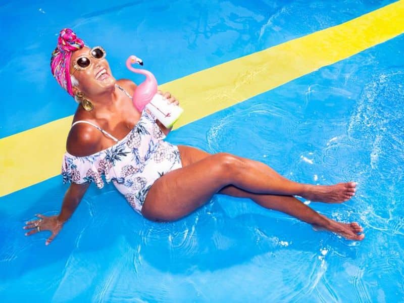 A woman lounging in the pool on a cruise ship, wearing a colorful headscarf and sunglasses, holding a drink with a pink flamingo floatie.