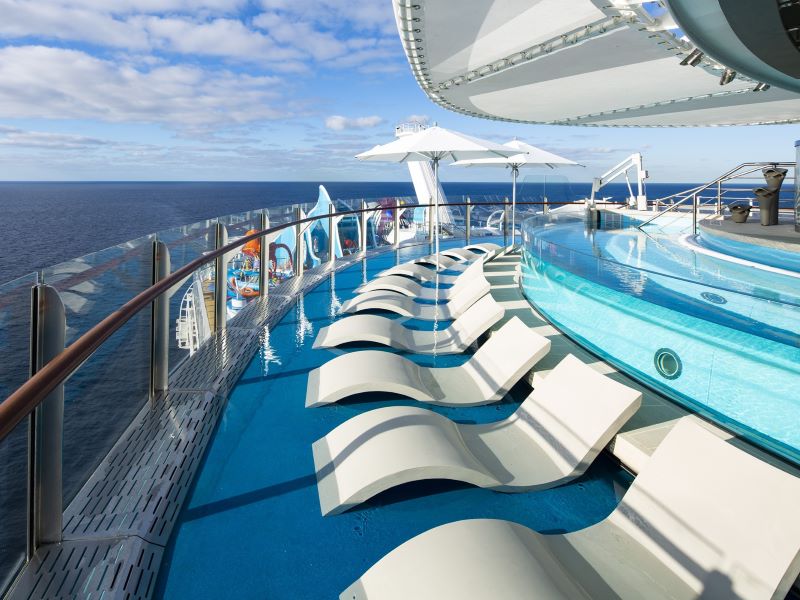 This image showcases the Suite Sun Deck on the Wonder of the Seas, the world's largest cruise ship, providing an exclusive retreat for suite guests. The deck features a modern, curved infinity pool that seems to merge with the ocean horizon. Sleek, white sun loungers are neatly arranged, offering passengers a comfortable place to relax and enjoy the panoramic sea views under large white umbrellas. The overall atmosphere is one of tranquility and luxury, set against the backdrop of a clear blue sky and open sea, emphasizing the premium experience aboard the ship.