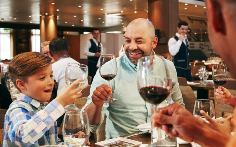 A joyful family moment at a restaurant with a young boy playfully toasting with water, mirroring a smiling bald man with a glass of red wine, amidst a bustling dining setting with attentive staff in the background.