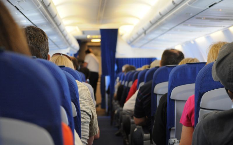 The image captures a perspective view inside an airplane cabin, focusing on the back of blue seats and passengers waiting or relaxing. In the aisle, a flight attendant can be seen attending to the passengers, and overhead compartments are slightly open. The setting conveys a typical scene during a flight, emphasizing the compact and communal experience of air travel.