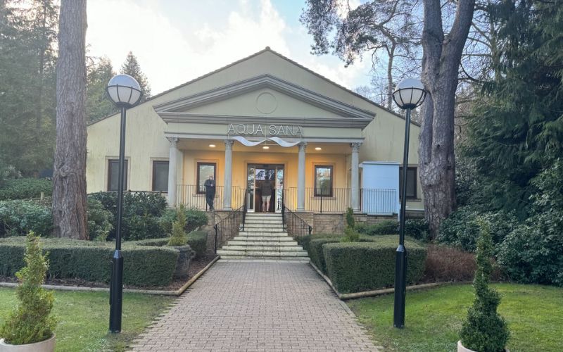 The entrance to Aqua Sana spa nestled among tall trees, featuring a classic building with a welcoming yellow façade, grand white columns, and the spa's name prominently displayed. The manicured pathway leading up to the spa suggests a serene and luxurious retreat.