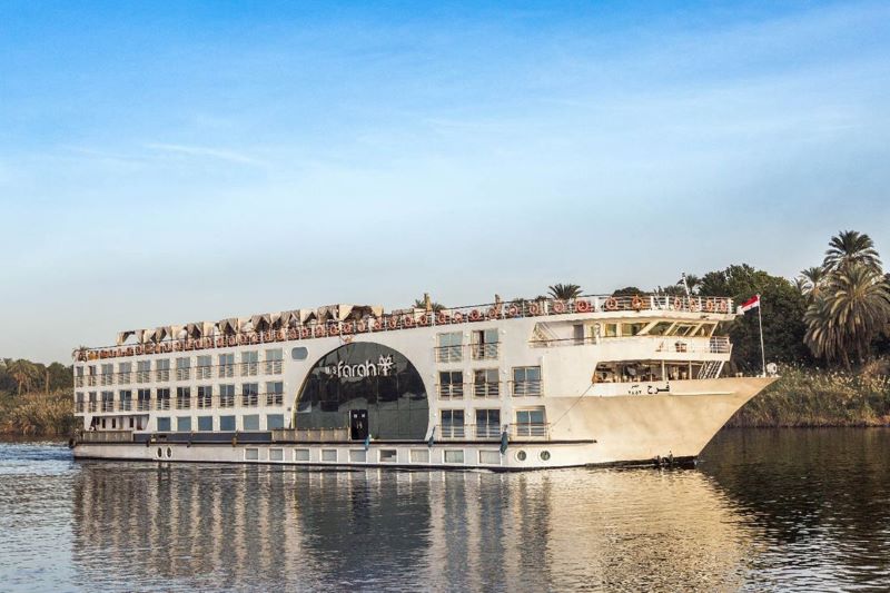 The Avalon Farah, a modern river cruise ship by Avalon Waterways, sails calmly on the Nile. Its distinctive round windows and streamlined design give it a contemporary look. Passengers enjoy the view from the upper deck, taking in the serene river and lush palm trees lining the bank, reflecting the unique blend of luxury and exploration that river cruises offer.