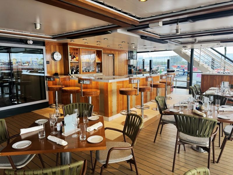 A sophisticated bar and restaurant area aboard the Seabourn Venture, featuring a warmly lit wooden bar counter lined with high stools and a dining area with elegantly set tables. The outdoor setting allows for natural light and views of the port, while the retractable roof overhead can adjust to various weather conditions. The combination of modern design and classic nautical elements creates a welcoming ambiance for guests to enjoy meals and drinks in an upscale, yet relaxed atmosphere.