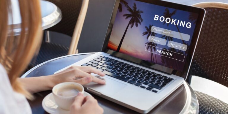 This image features a woman sitting at a café table using a laptop displaying a booking interface with options for 'Destination', 'Arrival', and 'Departure', set against a palm tree and sunset background. She is also holding a cup of coffee. The focus is on facilitating travel arrangements in a relaxed, outdoor setting.