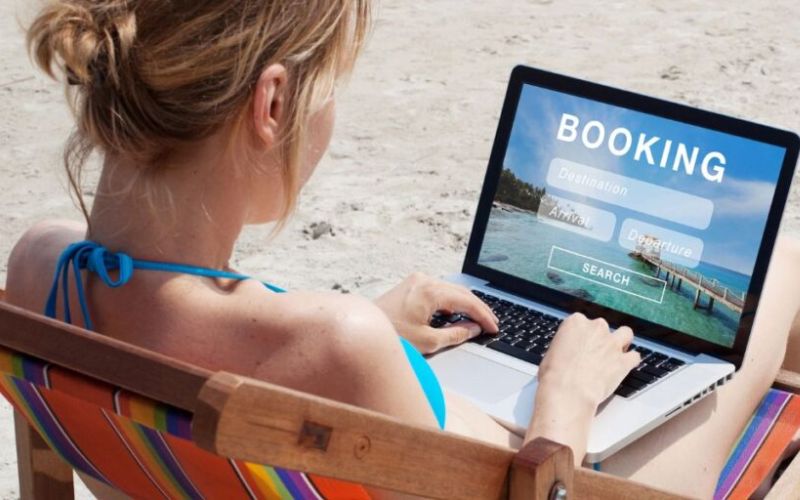 This image shows a woman from behind, seated on a colorful beach chair, using a laptop with a screen displaying a travel booking interface for destination, arrival, and departure options. She's on a sandy beach, symbolizing the ease of planning vacations even while enjoying a relaxed beach setting. The setup underscores the convenience of modern digital tools that enable holiday planning from any location.
