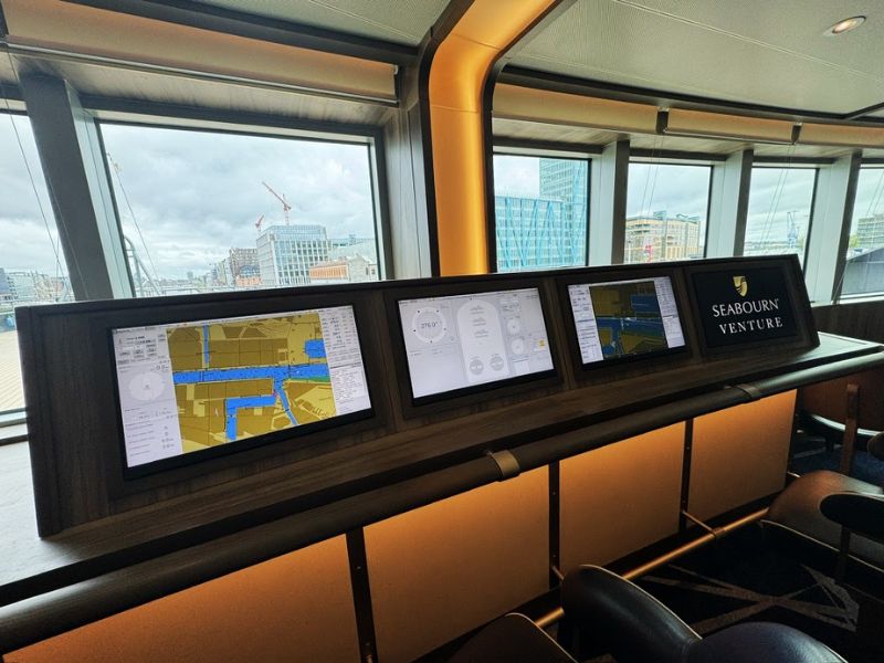 Sophisticated bow lounge on the Seabourn Venture, which features a modern informational setup. This area is equipped with multiple screens displaying navigational charts, ship statistics, and other data relevant to the cruise's current location and conditions. The lounge offers guests the opportunity to stay informed about their journey in real-time while enjoying the comfort of the venue. The panoramic windows provide ample natural light and a view of the ship's surroundings, enhancing the space's open and airy feel. This setup combines functionality with aesthetic appeal, making it a prime spot for relaxation and learning onboard.