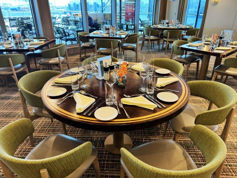 The buffet area on the Seabourn Venture is elegantly set up with a round wooden table at the forefront, surrounded by plush green chairs. The table is neatly laid with white plates, silverware, and cloth napkins accented with yellow, matching the cheerful orange flower centerpiece. In the background, similar tables are visible, creating a harmonious dining environment. The overall setting combines comfort with refined design, ideal for guests enjoying their meals while taking in the panoramic views from the large windows surrounding the dining area.