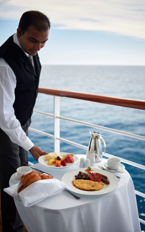 A well-dressed butler helping to serve a meal on a cruise ship terrace with a beautiful sea view