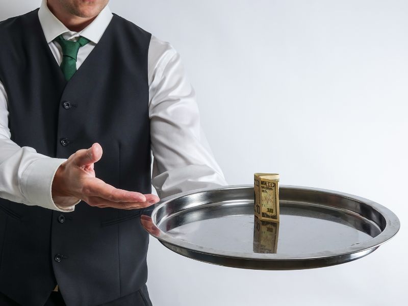 A waiter in a formal vest and tie presents a silver tray with a rolled banknote, symbolizing the discreet and appreciated gesture of tipping for excellent service.