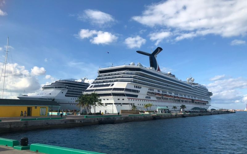 A Carnival cruise ship with its distinctive funnel docked at a tropical port, flanked by bright blue skies and calm sea waters, with a row of yachts and a colorful building to the left.