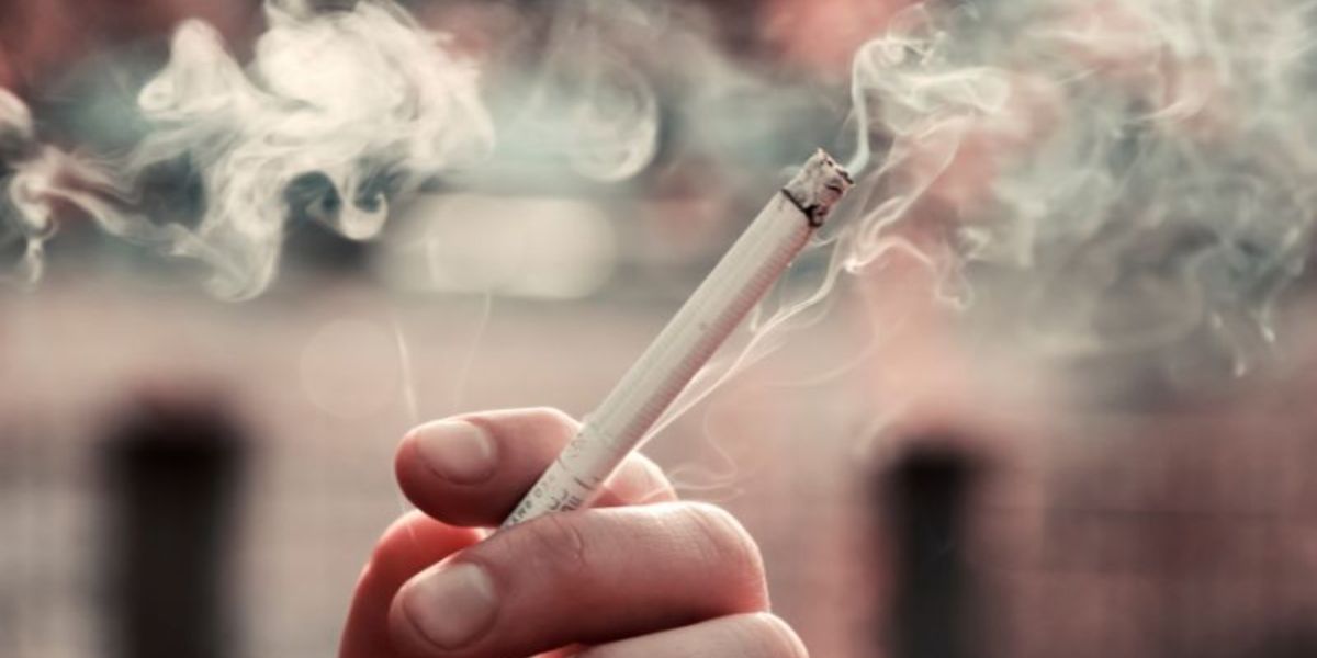 Close-up of a lit cigarette held between fingers, with smoke curling upwards, on the blurred background of a cruise ship deck.